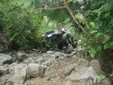 1985 Ford Bronco - A dark green Ford Bronco is navigating a rocky, overgrown trail, with its front tires angled upwards on large boulders. The vehicle is surr…