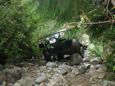 1985 Ford Bronco - A dark colored SUV is navigating a rocky, wooded trail. The vehicle is angled to the right, with its rear tires on large rocks.