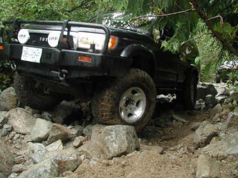 1985 Ford Bronco - A black off-road SUV navigates a rocky trail, with its front tire high on a boulder. The vehicle features a heavy-duty front bumper with au…