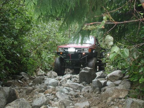 1985 Ford Bronco - A red 4x4 off-road vehicle navigates a rocky, wooded trail. The vehicle has large tires and a modified front bumper.