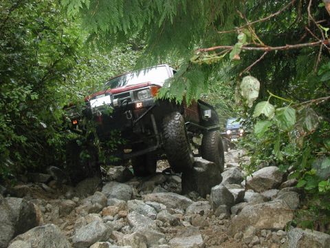 1985 Ford Bronco - A red off-road vehicle navigates a rocky, wooded trail, with another vehicle visible further down the path. The vehicle is tilted as it cli…