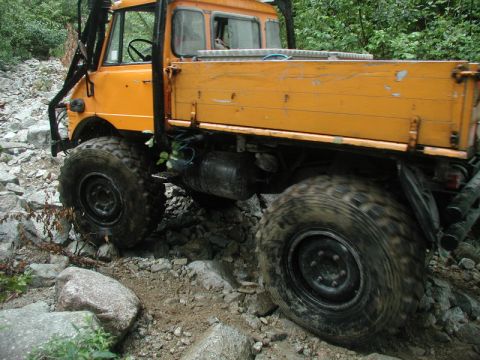 1985 Ford Bronco - A bright orange Unimog truck navigates a rocky, off-road trail with large, aggressive tires. The truck's bed is visible, showing wooden pla…