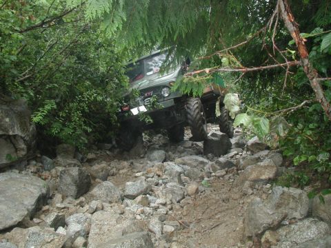 1985 Ford Bronco - A green off-road vehicle navigates a rocky, wooded trail, with its large tires gripping the uneven terrain. The vehicle appears to be a mod…