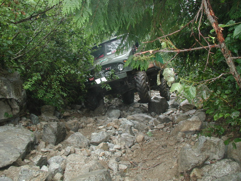 Green off-road vehicle driving over a rocky trail surrounded by trees.