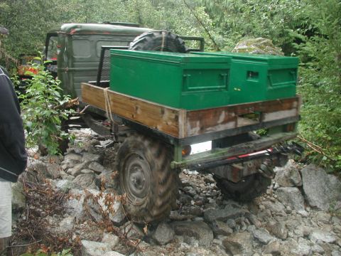 1985 Ford Bronco - A green military-style truck with a trailer carrying two green toolboxes navigates a rocky, wooded trail. The truck's large tires are desig…