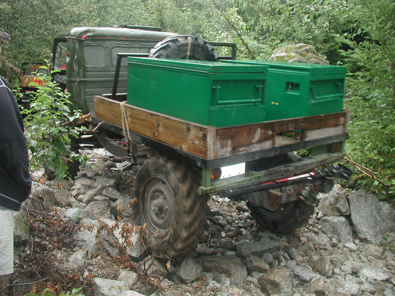 Green military truck with trailer and toolboxes on a rocky forest trail.