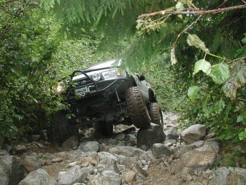 1985 Ford Bronco - A lifted Toyota 4Runner navigates a rocky, wooded trail, with its front tires climbing over large boulders.