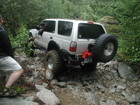 1985 Ford Bronco - A silver SUV with large off-road tires navigates a rocky trail, with a spare tire mounted on the rear carrier. A person stands to the left,…