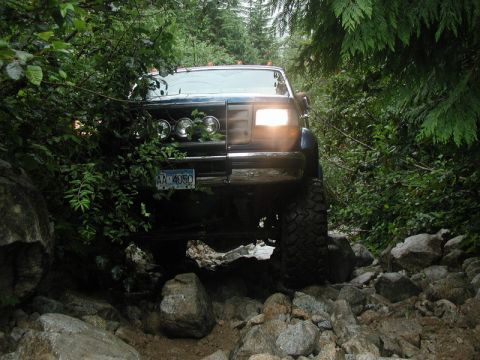 1985 Ford Bronco - A dark blue, lifted 1985 Ford Bronco navigates a rocky, wooded trail. The front of the vehicle is visible, with its headlights on and a lic…