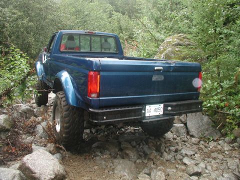 1985 Ford Bronco - The rear driver's side of a lifted, dark blue pickup truck is shown navigating a rocky, off-road trail. The truck has large, aggressive tir…