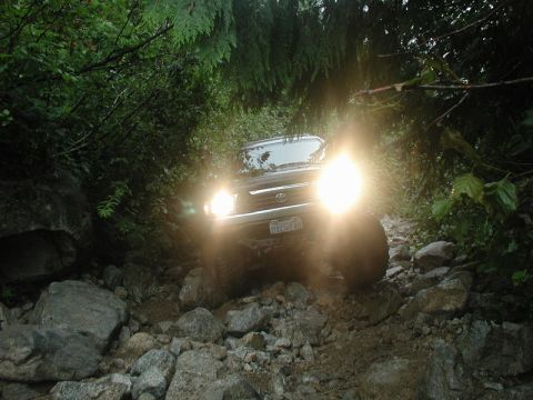 1985 Ford Bronco - A dark-colored SUV navigates a rocky, wooded trail with its headlights on. The vehicle is surrounded by large boulders and dense foliage.