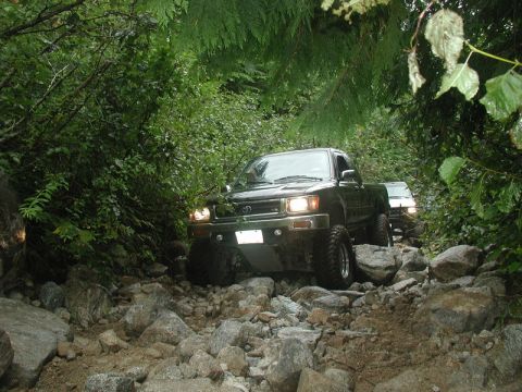 1985 Ford Bronco - Two dark-colored pickup trucks navigate a rocky, wooded trail. The lead truck is a Toyota pickup with its headlights on, driving over large…
