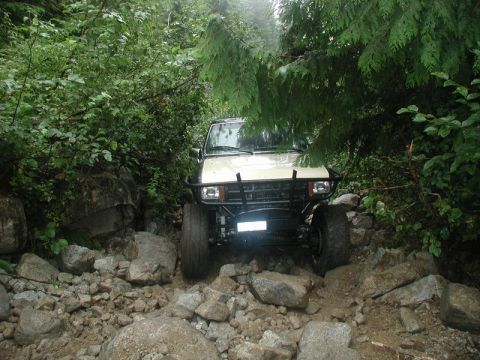 1985 Ford Bronco - A tan off-road vehicle navigates a rocky, wooded trail, with its front bumper and tires visible.