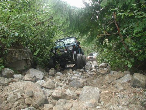 1985 Ford Bronco - A dark green Suzuki Samurai navigates a rocky, wooded trail. Another off-road vehicle is visible further down the path.