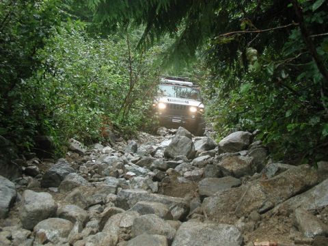 1985 Ford Bronco - A white Hummer H1 navigates a very rocky, overgrown trail in a forest setting. The vehicle's headlights are on as it moves through the chal…
