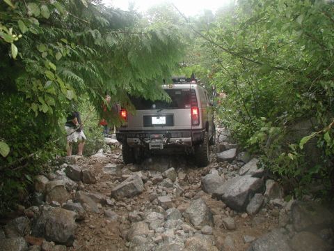 1985 Ford Bronco - A silver Hummer H2 navigates a rocky, overgrown trail with several people standing nearby. The vehicle is viewed from the rear as it moves…