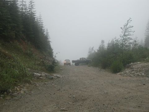 1985 Ford Bronco - A dark colored SUV and a pickup truck drive on a gravel road surrounded by trees and fog.