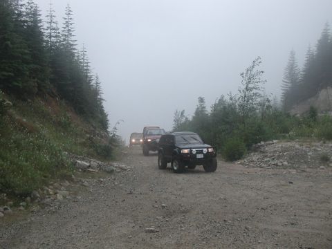 1985 Ford Bronco - A line of four-wheel drive vehicles, including a black SUV in the foreground, navigate a foggy, gravel mountain road lined with evergreen t…