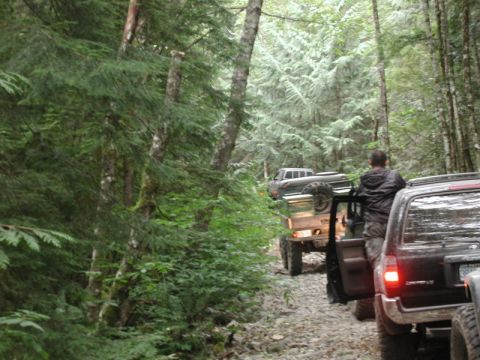 1985 Ford Bronco - A line of off-road vehicles drives down a gravelly, wooded trail. The lead vehicle appears to be a dark-colored SUV with its driver's side…