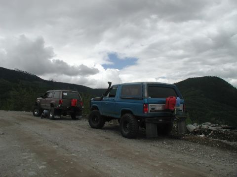 1985 Ford Bronco - Two modified off-road SUVs are parked on a gravel road with mountains in the background under a cloudy sky. The blue SUV in the foreground…