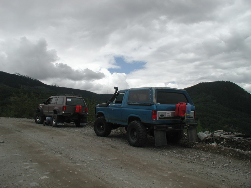 Two lifted SUVs on a gravel road with mountains and clouds.