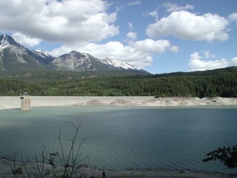 1985 Ford Bronco - A large dam holds back a reservoir of water, with a dense evergreen forest and snow-capped mountains in the background under a partly cloud…