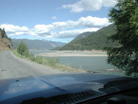 1985 Ford Bronco - The hood and windshield of a blue vehicle are visible in the foreground, looking out at a scenic mountain landscape with a large body of wa…