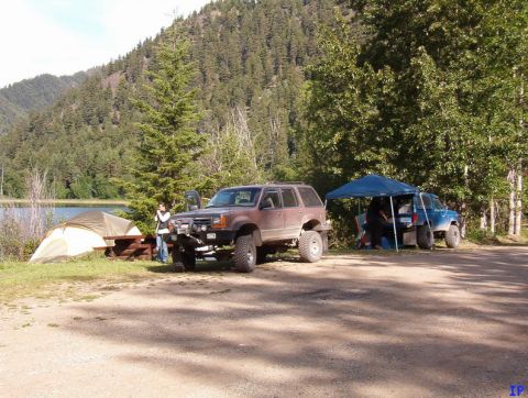 1985 Ford Bronco - A campsite scene features two modified off-road vehicles, a brown SUV and a blue pickup truck, parked near a lake and a wooded hillside. A…