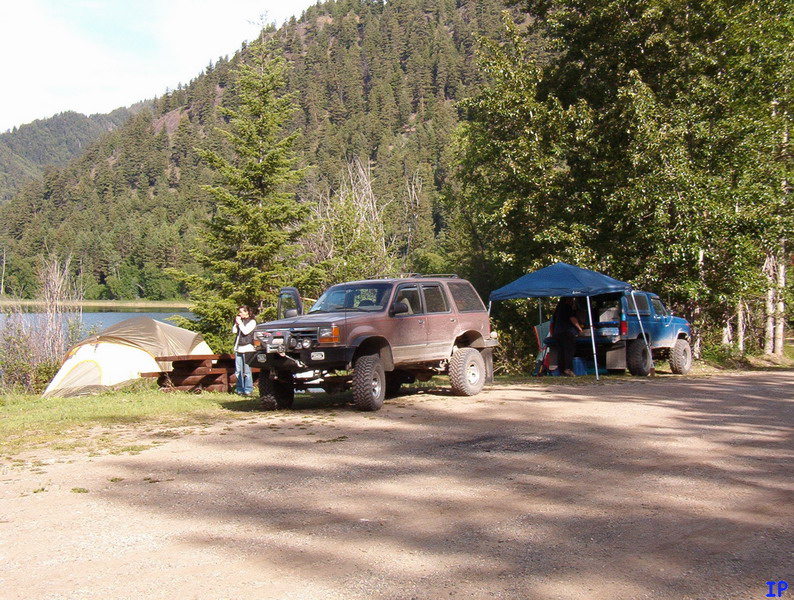 Two modified off-road vehicles at a campsite near a lake.