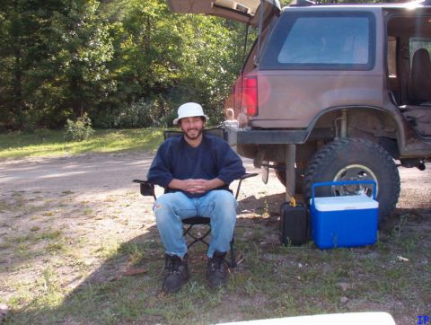 1985 Ford Bronco - A man wearing a white bucket hat and jeans sits in a camping chair next to a lifted, muddy SUV with its rear hatch open. A blue cooler and…