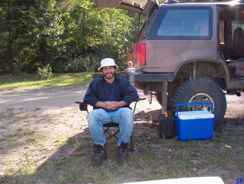 Man in camping chair next to lifted, muddy SUV with open hatch, cooler, and fuel can.