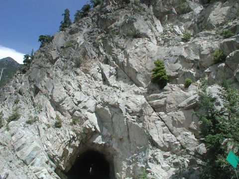 1985 Ford Bronco - A dark tunnel entrance is visible in the lower center of the image, carved into a steep, rocky mountainside. The surrounding rock face is r…