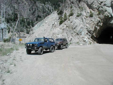 1985 Ford Bronco - A lifted blue 1980s Ford Bronco is parked on a dirt road next to a tunnel entrance in a mountainous area. A second SUV is parked behind it,…