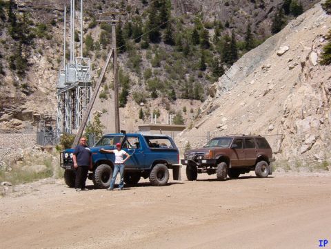 1985 Ford Bronco - Two off-road modified SUVs are parked on a dirt road in front of a rocky hillside with a power substation in the background. A blue Ford Br…