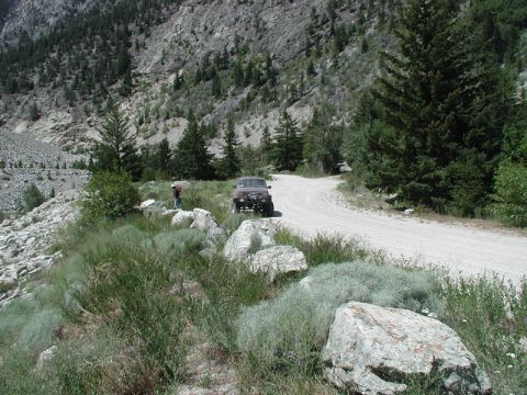 1985 Ford Bronco - A dark colored SUV is parked on a dirt road next to a rocky hillside covered with sparse vegetation and trees. A person wearing a backpack…