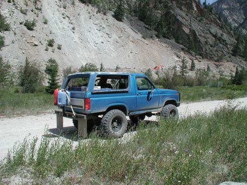 1985 Ford Bronco - A lifted blue 1985 Ford Bronco is parked on a dirt road in a mountainous, rocky area with sparse vegetation. The rear of the vehicle shows…