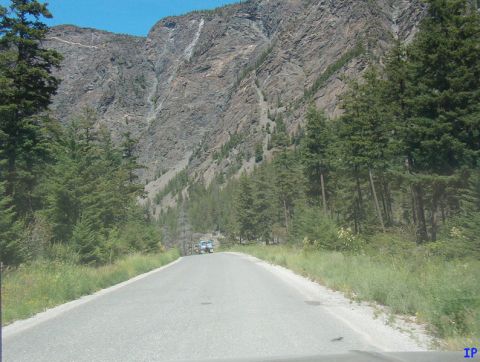 1985 Ford Bronco - A light blue SUV is driving on a paved road through a mountainous area with pine trees and steep rock faces. The road curves to the right a…