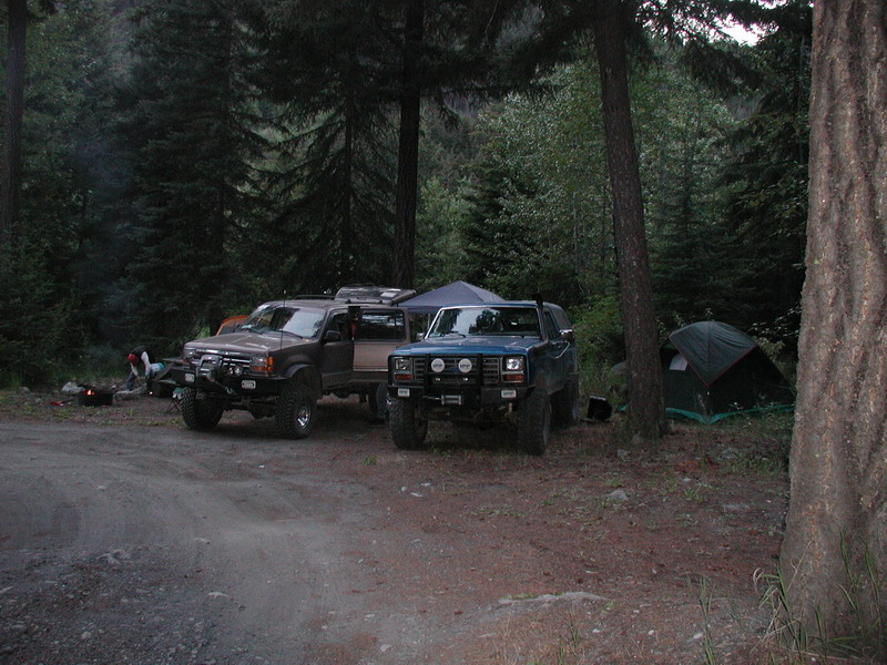 Two off-road trucks parked at a campsite in a forest with a tent and campfire.