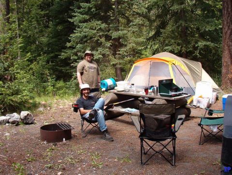 Two men camping next to a tent and picnic table in a forest.