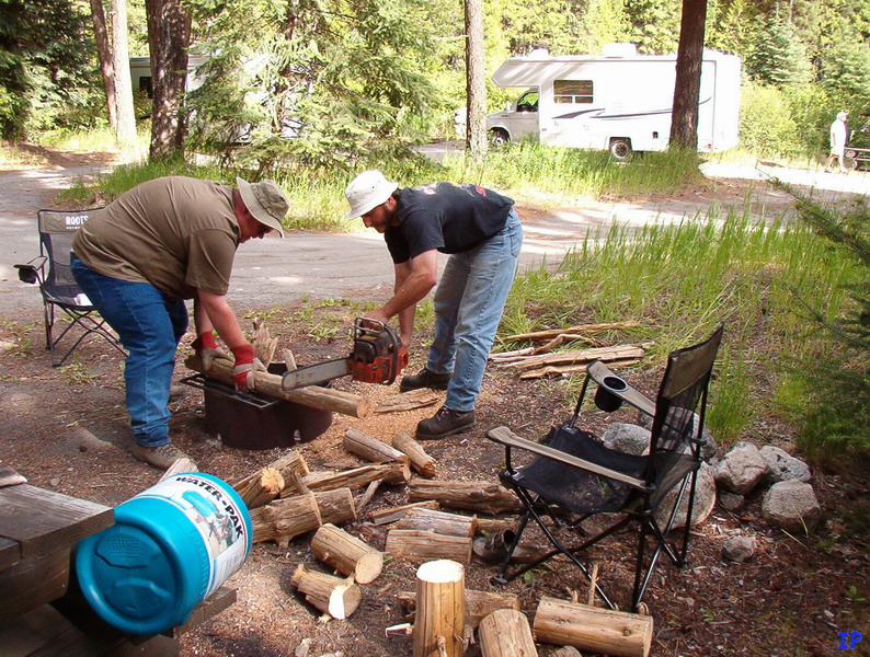 Two men cutting logs with a chainsaw at a campsite with an RV in the background.