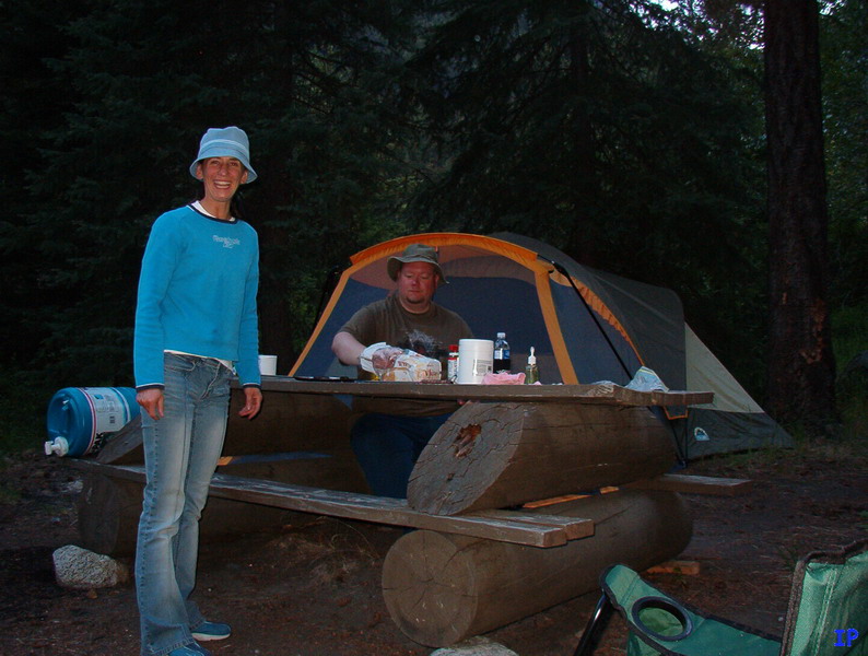 A woman and a man camping next to a tent and a log picnic table.
