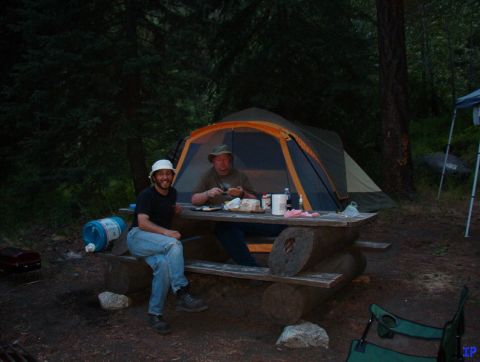Two men at a picnic table in a campsite with a tent.