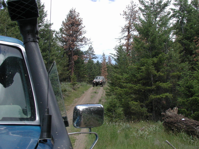 Blue SUV with snorkel driving on a dirt road in a forest, with another SUV ahead.