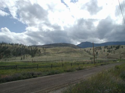 Rural landscape with rolling hills, scattered trees, a dirt road, and a cloudy sky.