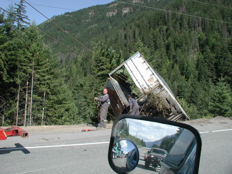 Two men securing a tipped-over semi-trailer with cables on the side of a highway.