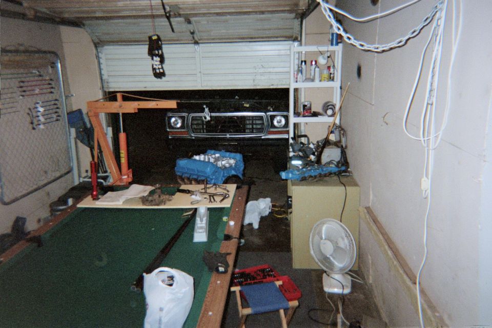 Garage with Bronco front end at door, blue engine block on floor, engine hoist, and parts on a pool table