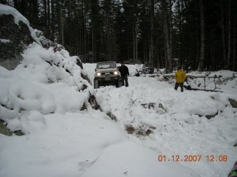 White 1985 Ford Bronco driving on a snowy, wooded trail with two people standing nearby.
