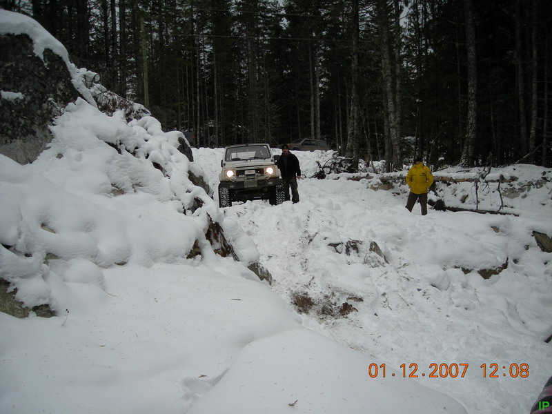 White 1985 Ford Bronco driving on a snowy, wooded trail with two people standing nearby.