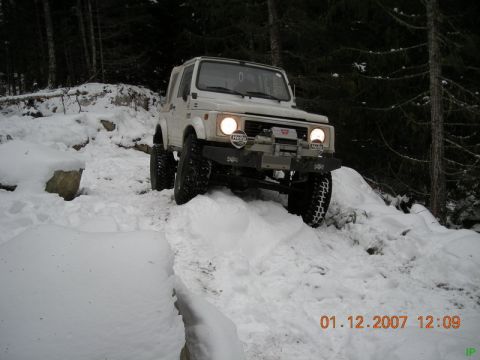 White Suzuki Samurai driving up a snowy forest hill.
