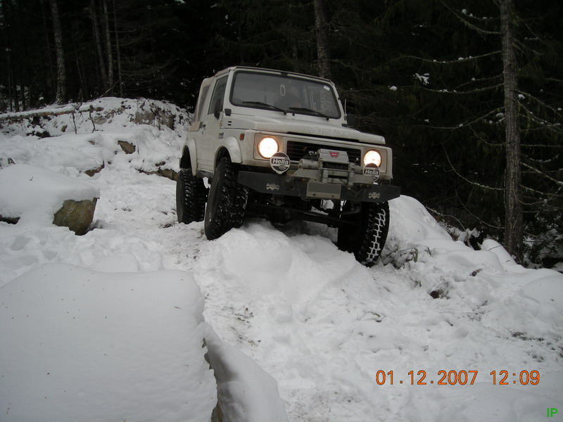 White Suzuki Samurai driving up a snowy forest hill.